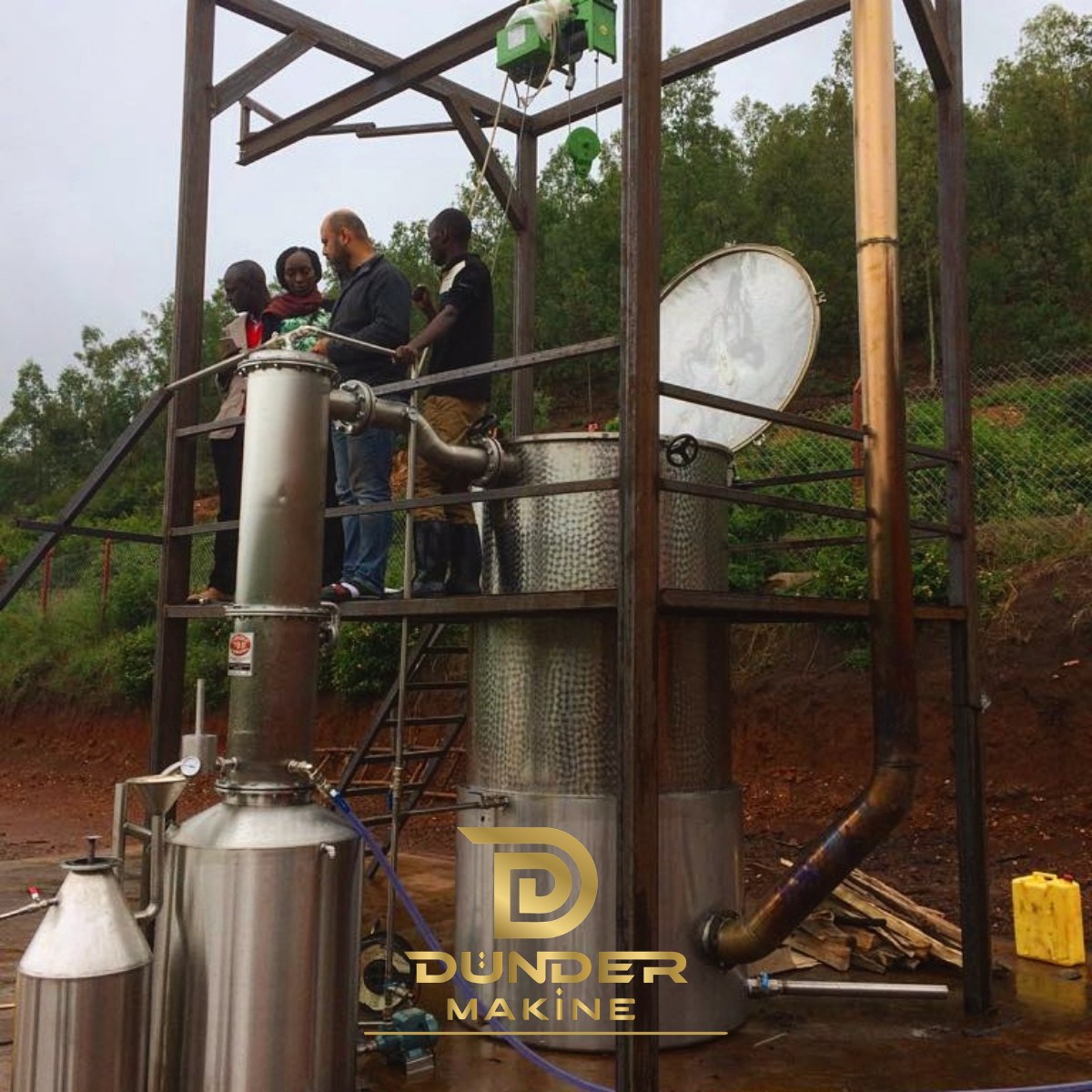Wood-fired distillation unit with basket hoist, condenser and separator during start-up on a hillside site